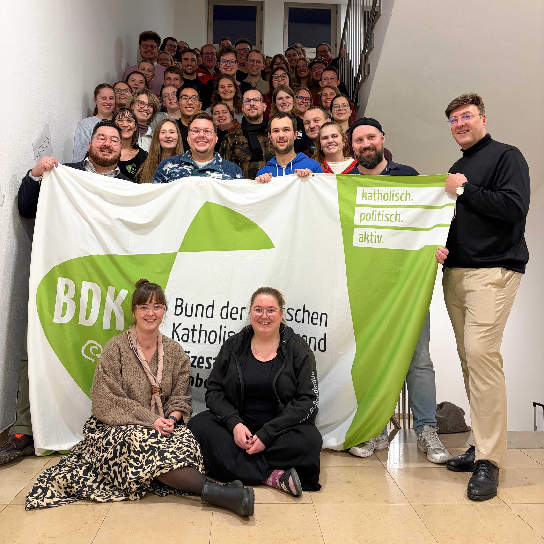 Gruppenbild von der Diözesanversammlung in einem Treppenhaus. Einige Personen halten ein großes Banner mit dem Logo des BDKJ.