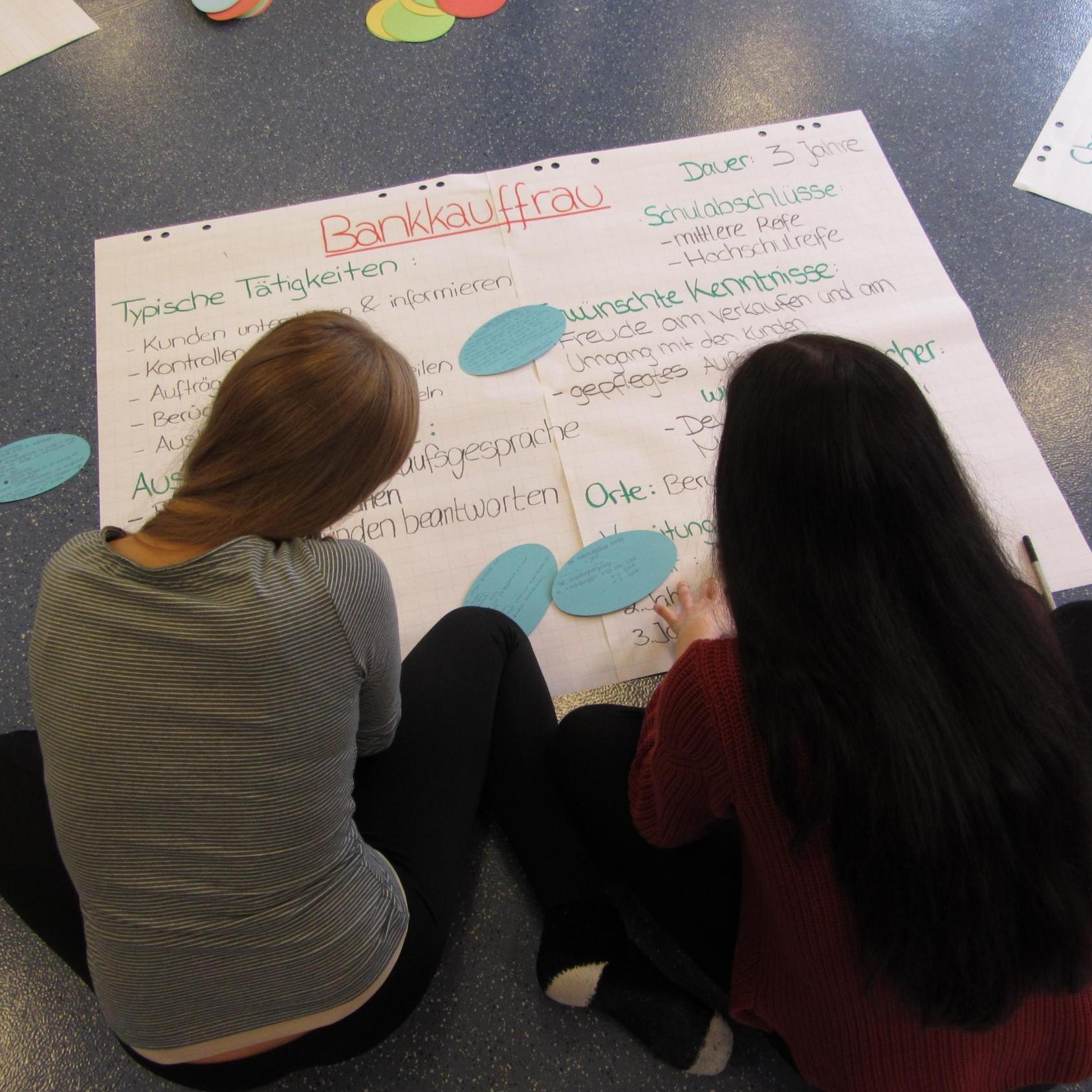 Zwei Jugendliche betrachten ein auf dem Boden liegendes Plakat mit Informationen zum Beruf der Bankkauffrau.