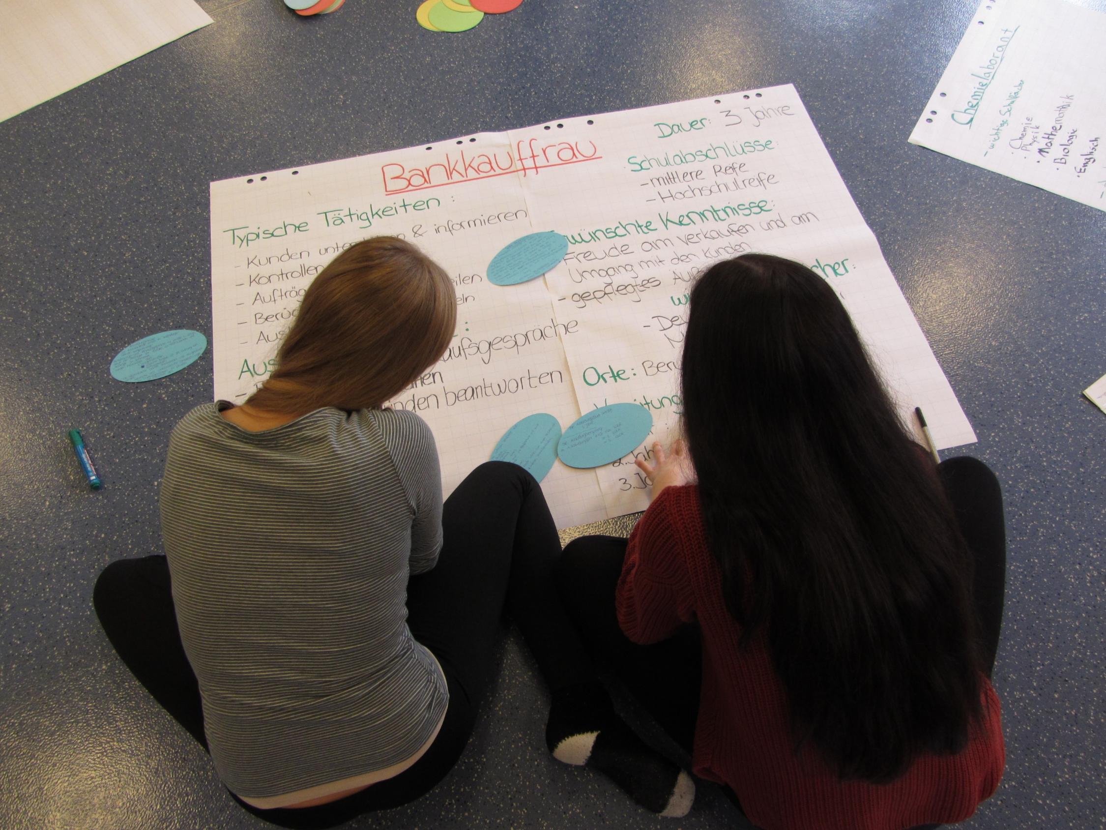 Zwei Jugendliche betrachten ein auf dem Boden liegendes Plakat mit Informationen zum Beruf der Bankkauffrau.