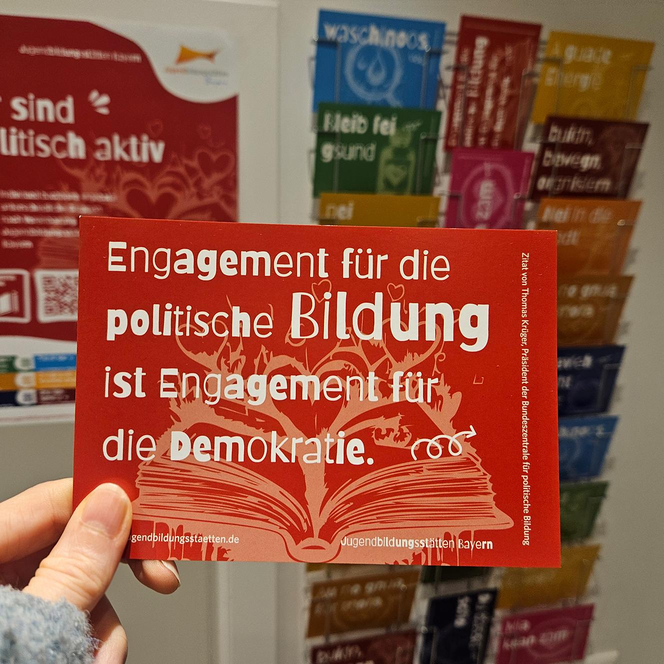 Vor einem unscharfen Ständer mit verschiedenen Postkarten im Hintergrund wird eine Postkarte mit der Aufschrift 'Engagement für die politische Bildung ist Engagement für die Demokratie' ins Bild gehalten.