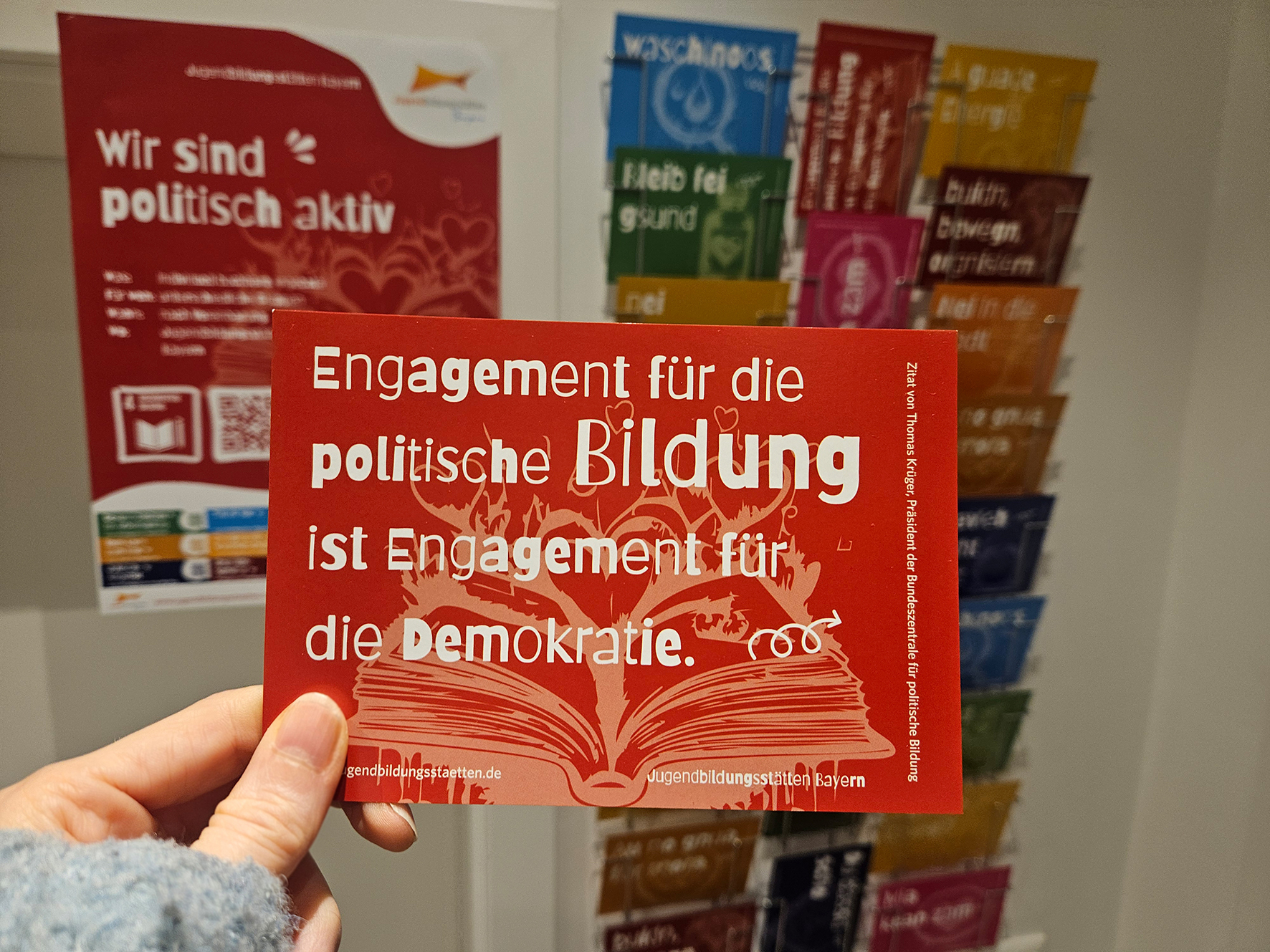 Vor einem unscharfen Ständer mit verschiedenen Postkarten im Hintergrund wird eine Postkarte mit der Aufschrift 'Engagement für die politische Bildung ist Engagement für die Demokratie' ins Bild gehalten.